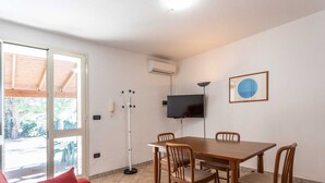 Ein Zimmer mit einem Holzesstisch, vier Stühlen, einem roten Sofa, einem an der Wand montierten Fernseher und einer Klimaanlage.