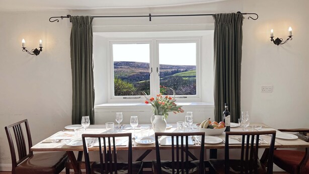 A dining area with a table set for six, a window with a view of rolling hills, and two wall-mounted light fixtures. - Brightworthy Farm, Withypool on Exmoor (Withypool)