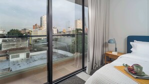Hotellihuone, jossa on suuri ikkuna, valkoisilla lakanoiden peitetty sänky, yöpöytä lampun kanssa ja tarjotinalusta, jossa on hedelmä ja kuppi.