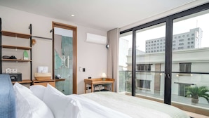 Hotellihuone, jossa on suuri sänky, työpöytä lampun kanssa, hylly, jossa on hygieniatuotteita, ja suuri ikkuna, josta avautuu näkymä rakennuksiin.