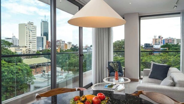 A modern hotel room with a balcony, a round table with fruit, a sofa, and a large window offering a city view.