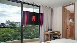 A modern hotel room with a large window offering a city view, a flat-screen TV mounted on the wall, a wooden desk with a chair, and a bed with a white comforter.