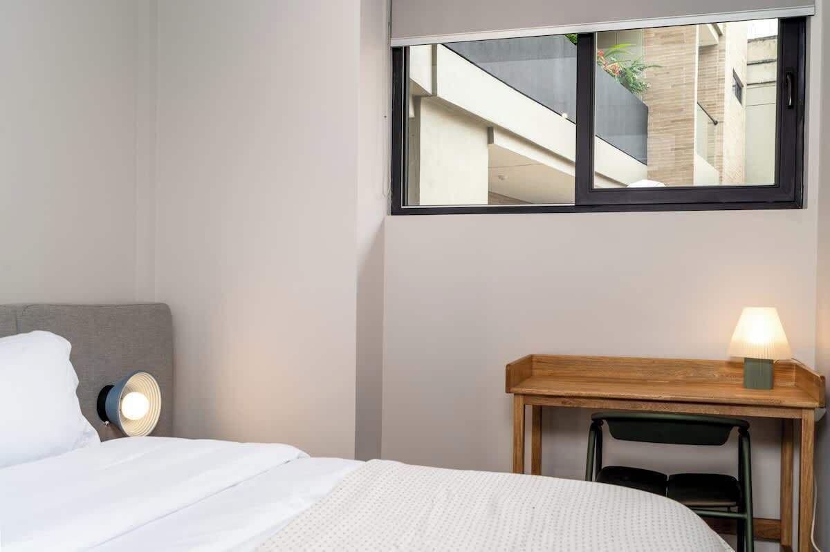 Ein Hotelzimmer mit einem Bett, einer Nachttischlampe, einem kleinen Holztisch mit Lampe und einem Fenster mit Blick auf ein Gebäude und Grünflächen.