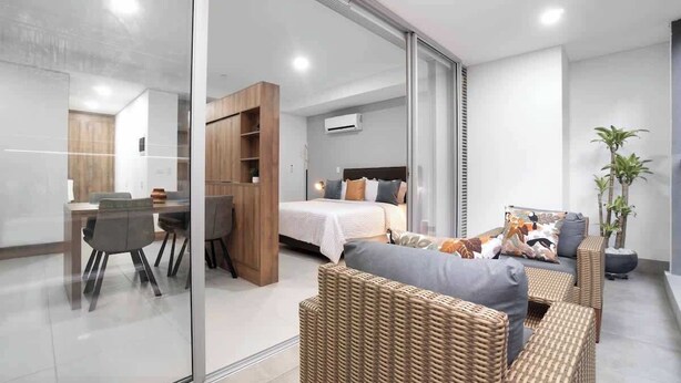 Una habitación de hotel moderna con un separador de vidrio que divide la sala de estar del dormitorio. La habitación cuenta con una cama grande, una mesa de comedor con sillas y un sofá de mimbre.