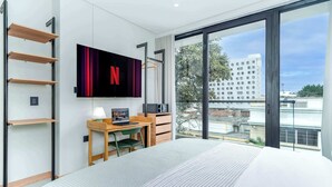 Moderni hotellihuone, jossa on suuri sänky, työpöytä kannettavalla tietokoneella, seinälle asennettu televisio ja parveke, jolta on näkymä kaupunkiin.