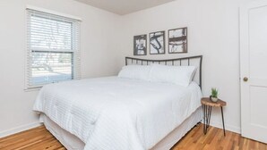 A bedroom with a bed, a window with blinds, a door, and three framed pictures on the wall. - Perfect 2-bedroom apartment near McKennan Park and Downtown! (Sioux Falls)