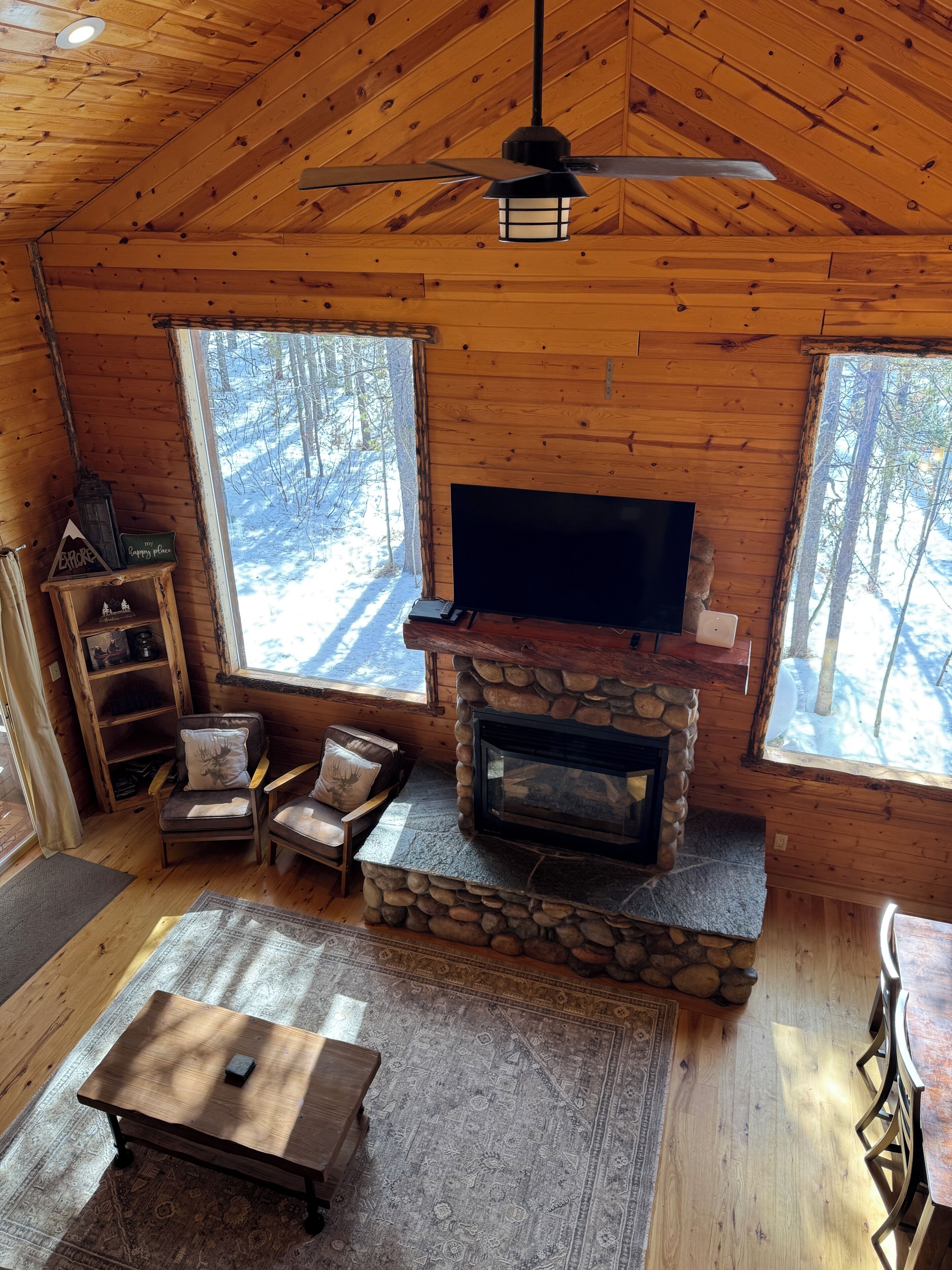 A cozy cabin interior with a stone fireplace, a television, wooden walls, and large windows overlooking a snowy forest.