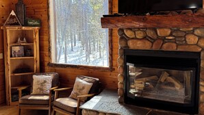 L’intérieur d’un chalet accueillant, avec une cheminée en pierre, des murs en bois, un téléviseur et une grande fenêtre donnant sur une forêt.
