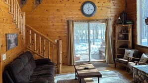 L’intérieur d’une cabane en rondins, avec un escalier en bois, un canapé en cuir noir, une table basse en bois et une grande fenêtre donnant sur un paysage enneigé.