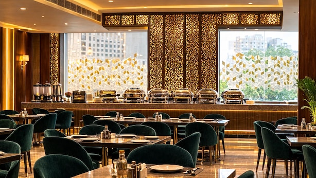 A restaurant with tables and chairs, a buffet area, and a city view through large windows. - Hotel RS (Agra)