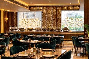 A restaurant with tables and chairs, a buffet area, and a city view through large windows. - Hotel RS  (Agra)