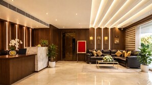 A modern lobby with a reception desk, leather sofas, potted plants, and a red accent wall. - Hotel RS (Agra)