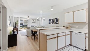 A modern kitchen with white cabinetry, a central island, and a dishwasher. There is a dining area with a table and chairs, and a living room with a sofa and a coffee table. - Walk in NO STAIRS 2 Bedroom Condo in Pointe Royale Golf Village NEW FOR 2026 (Branson)