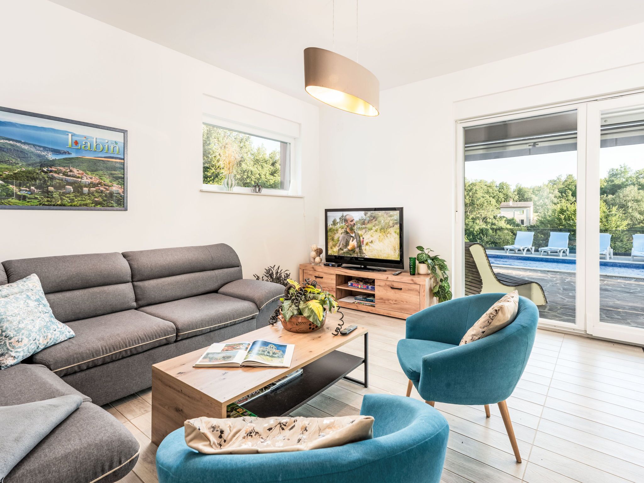 A modern living room with a sofa, a coffee table, a TV, and a view of a swimming pool.