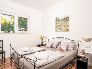 Een slaapkamer met een bed, twee nachtkastjes, een stoel, een raam met planten en een schilderij aan de muur.