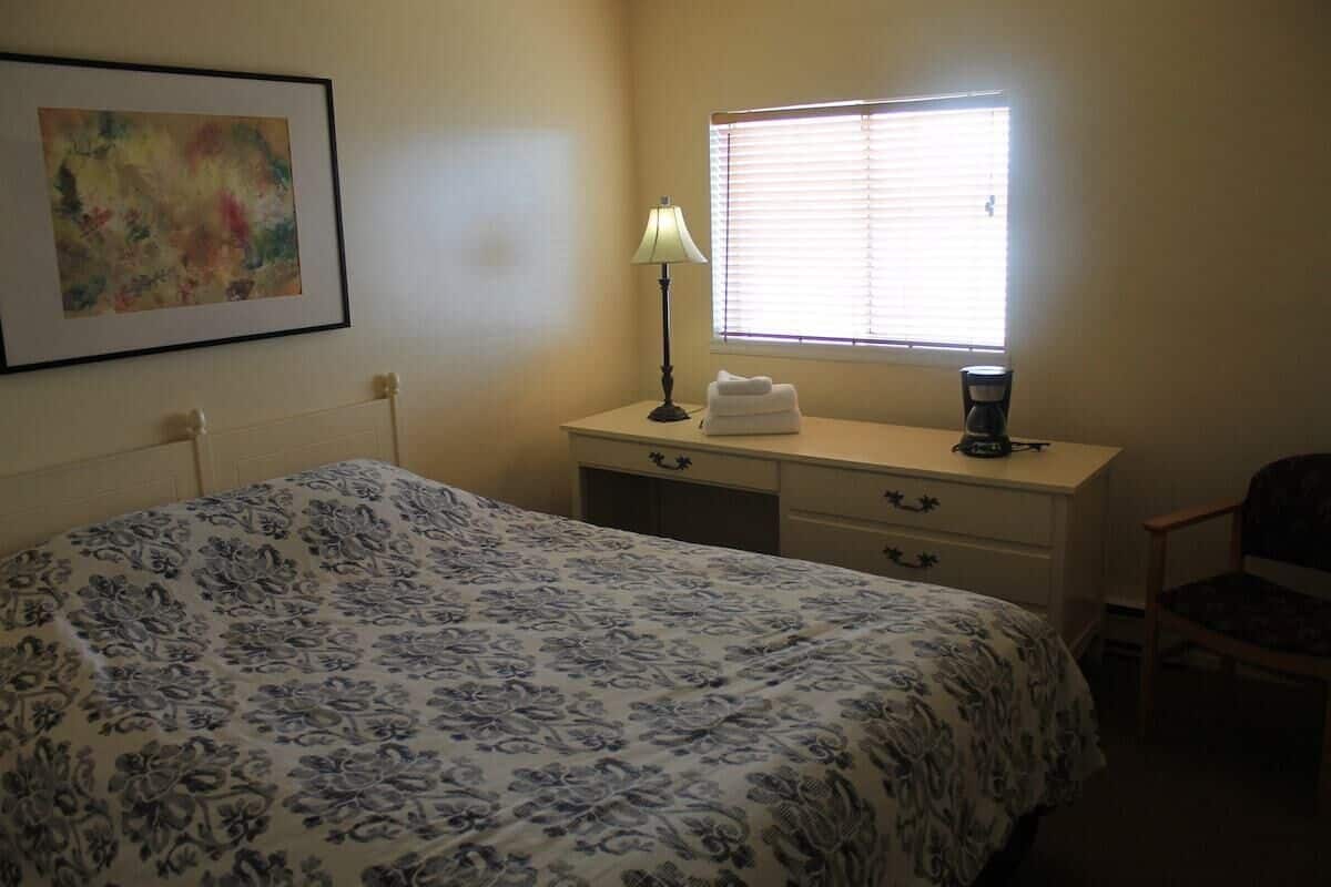 A bedroom with a bed, a bedside table with a lamp, a window with blinds, and a chair.
