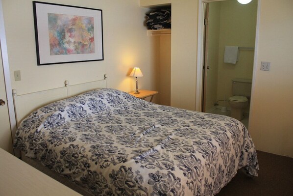 A bedroom with a bed, a bedside table with a lamp, a framed picture on the wall, and a bathroom visible through an open door.