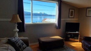 Una sala de estar con sofá, reposapiés, mueble para televisión y ventana con vistas a un lago y montañas.