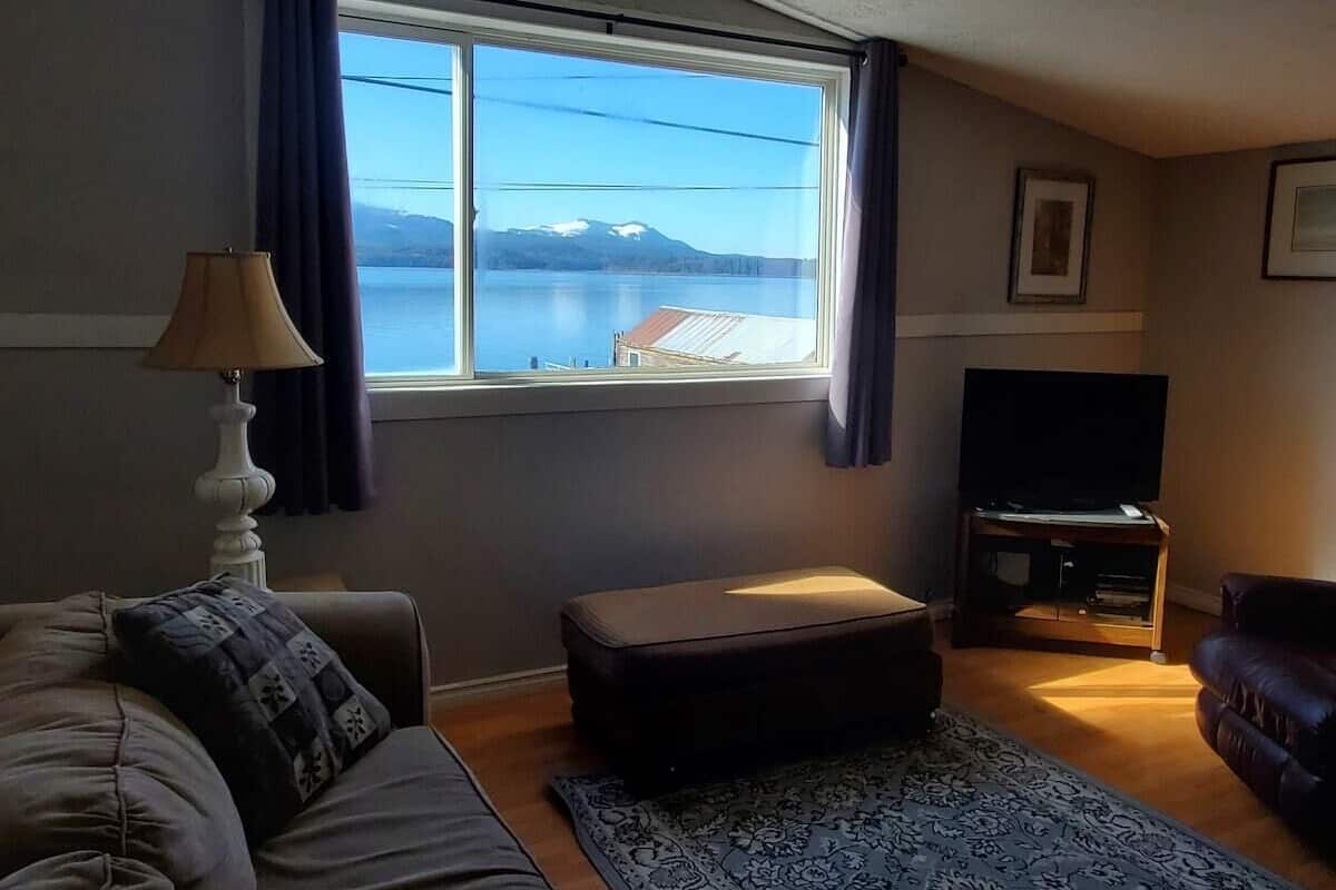 A living room with a sofa, ottoman, TV stand, and a window with a view of a lake and mountains.