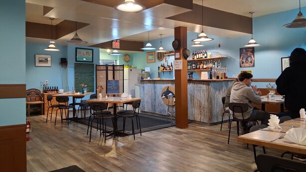 Un interior de restaurante con mesas y sillas de madera, lámparas colgantes y una zona de bar con botellas y vasos.