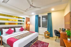 A hotel room with a bed, a desk, a chair, a window with curtains, a ceiling fan, and a wall with a decorative panel. - Hotel All Time Residency New Delhi (New Delhi)