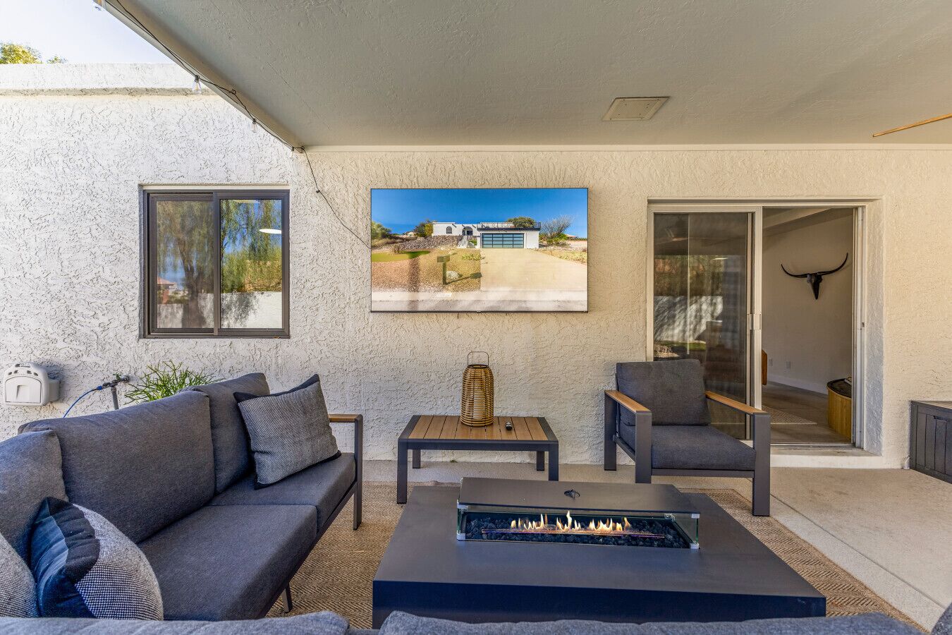 A patio with a gray sofa, a small table, a fire feature, and a view of a building through a window.