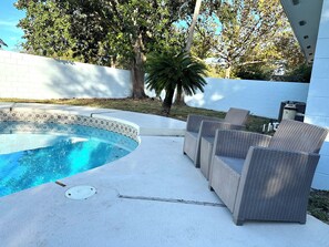 Ein Pool mit Fliesenrand, umgeben von einer Betonplattform. Im Hintergrund sind Korbsessel und eine Palme zu sehen.
