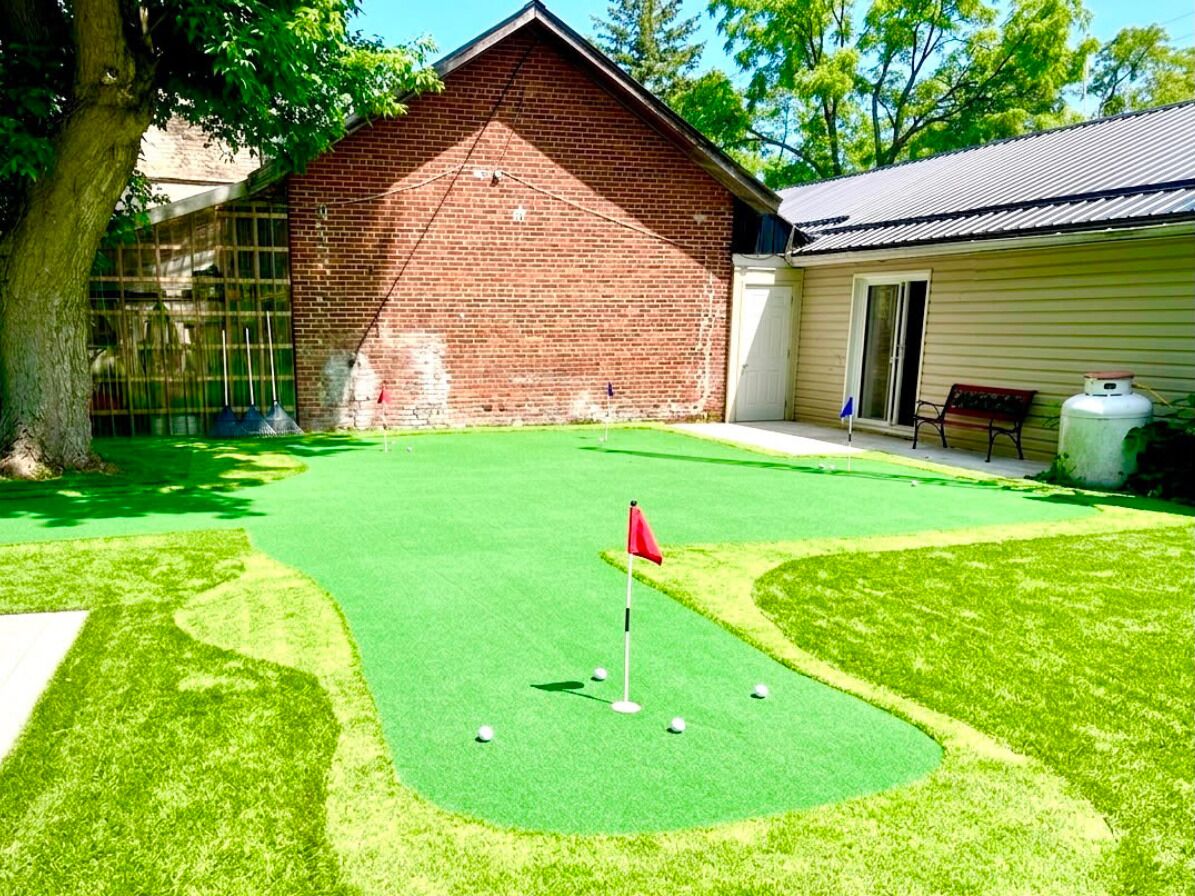 Ein Golfplatz mit künstlichem Rasen, eine rote Fahne, ein Backsteinhaus und ein Gebäude mit Schrägdach.