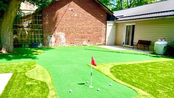 Ein Golfplatz mit künstlichem Rasen, eine rote Fahne, ein Backsteinhaus und ein Gebäude mit Schrägdach.