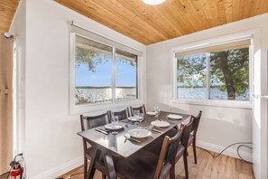 A dining area with a table set for four, large windows with a view of a lake, and a wooden ceiling. - #8 Cottage - Chile Lakefront (Hamilton)