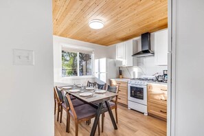 A modern kitchen with a wooden ceiling, white cabinets, a stove, and a dining area with chairs and a table set for four. - #8 Cottage - Chile Lakefront (Hamilton)