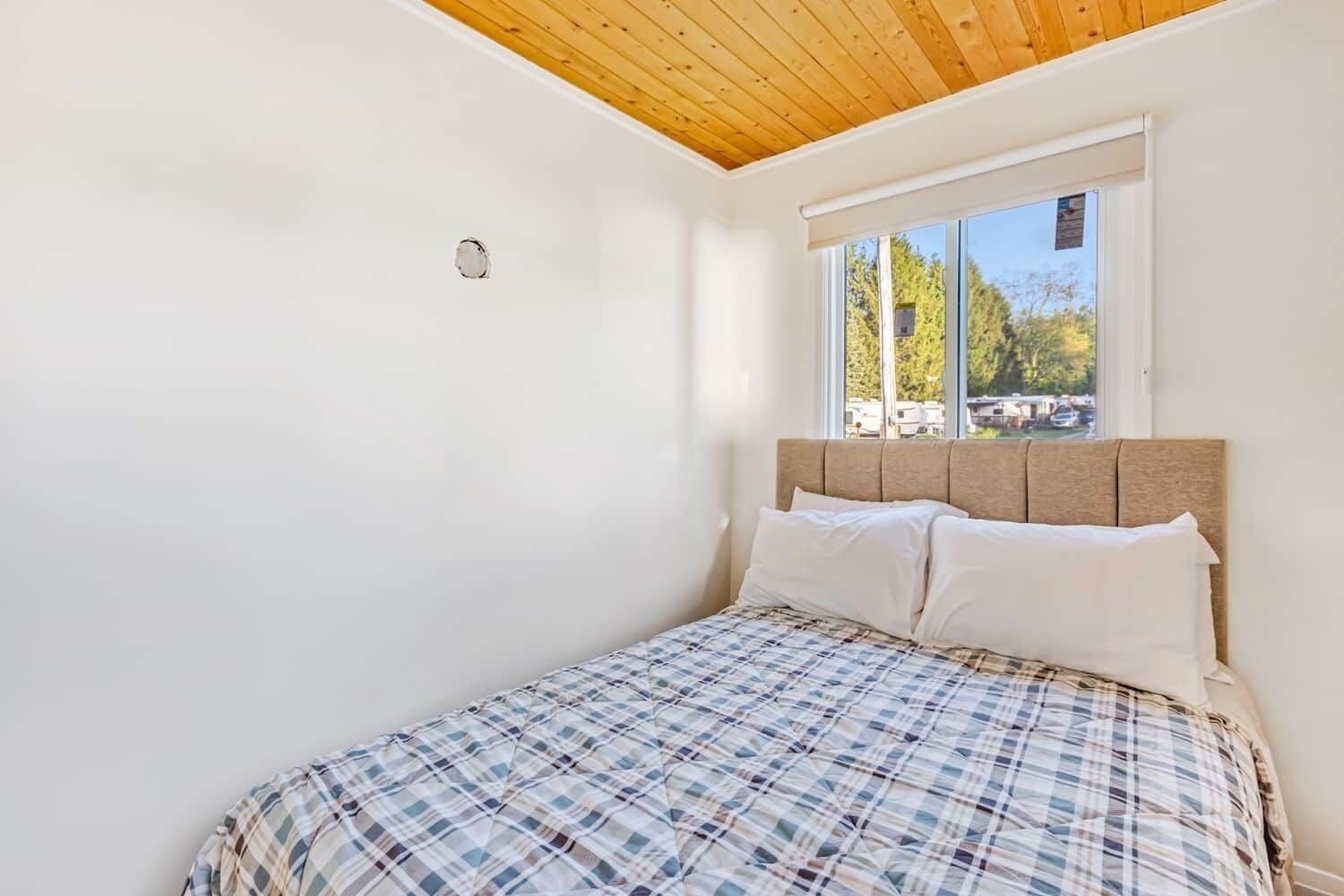 A bedroom with a wooden ceiling, a window with a view of trees, and a bed with a plaid bedspread.