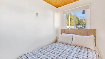 A bedroom with a wooden ceiling, a window with a view of trees, and a bed with a plaid bedspread.