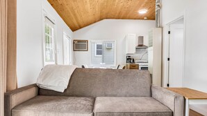 A cozy room with a sofa, a wooden ceiling, a kitchen area, and a window.