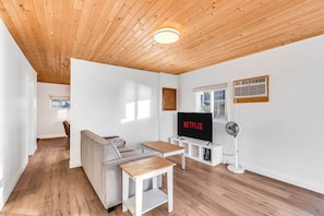 Ein modernes Wohnzimmer mit Holzbalkendecke, einer Couch, einem Couchtisch, einem Fernseher, der 'NETFLIX' zeigt, und einem Ventilator.