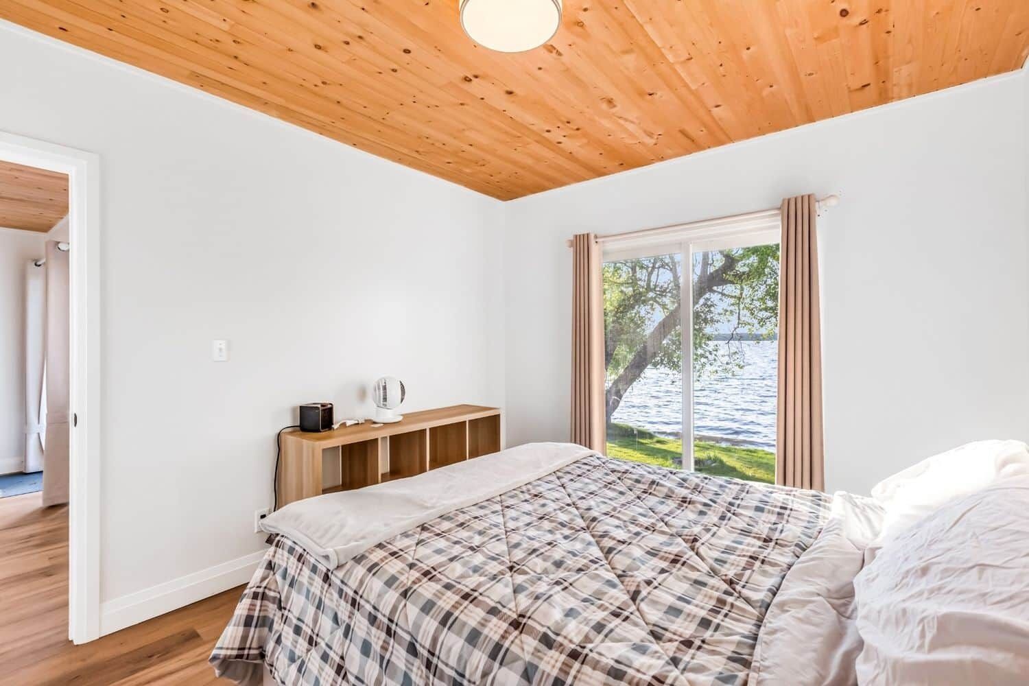 A bedroom with a wooden ceiling, a bed with a plaid bedspread, a bedside table with a lamp, and a window with a view of a lake.