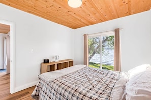 Ein Schlafzimmer mit Holzbalkendecke, einem Bett mit kariertem Überwurf, einem Nachttisch mit Lampe und einem Fenster mit Seeblick.