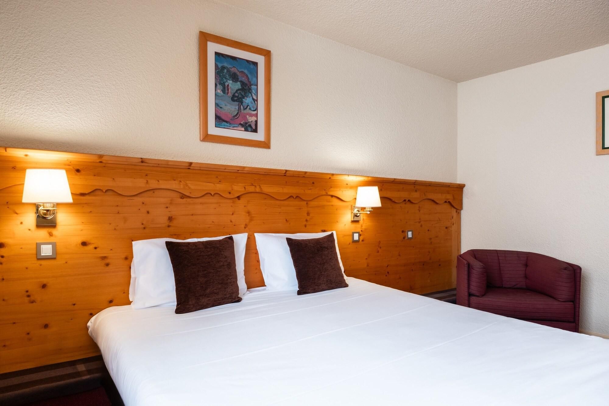 A hotel room with a wooden headboard, two bedside lamps, a bed with white linens and brown pillows, a red armchair, and a framed painting on the wall.