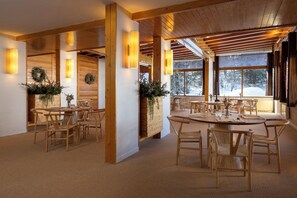 En inredning med trädetaljer, flera matbord och stolar, stora fönster med utsikt över ett snötäckt landskap och en varm belysning.