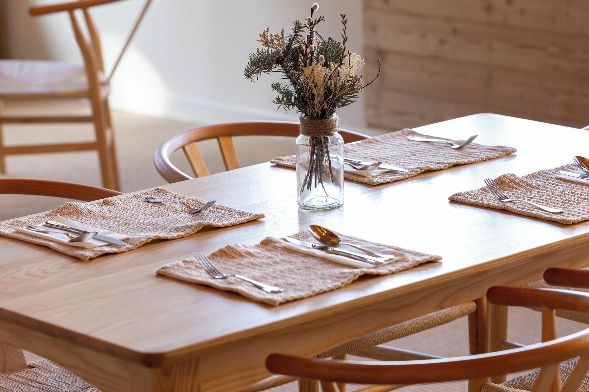 Un tavolo da pranzo in legno apparecchiato per quattro persone, con un sottopiatti intrecciato, posate e un vaso di vetro con fiori secchi per ogni posto.