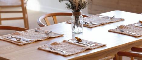 Un tavolo da pranzo in legno apparecchiato per quattro persone, con un sottopiatti intrecciato, posate e un vaso di vetro con fiori secchi per ogni posto.