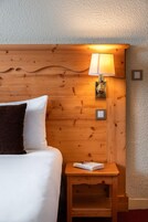 Une tête de lit en bois, une lampe de chevet, une petite table de chevet en bois avec un livre, et un lit blanc avec un oreiller foncé.