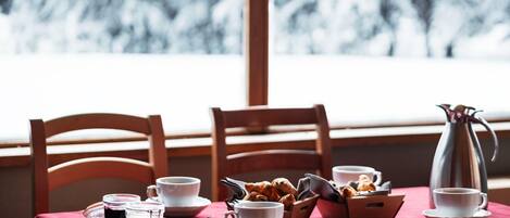 Een ontbijttafel met kopjes, een karaf en gebak, bij een raam met uitzicht op sneeuw.
