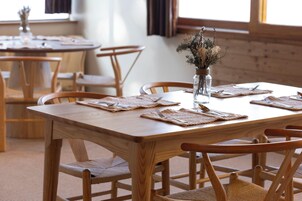 Un coin repas avec des tables et des chaises en bois, un vase contenant des fleurs séchées et une fenêtre avec des rideaux.