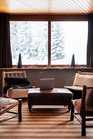 A cozy room with a wooden ceiling, a large window with a view of snow-covered trees, a laptop on a wooden ottoman, and two chairs with fur throw pillows. - Crystal Hôtel Courchevel (Les Allues)