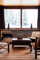 Une pièce chaleureuse avec un plafond en bois, une grande fenêtre donnant sur des arbres couverts de neige, un ordinateur portable posé sur un pouf en bois, et deux fauteuils agrémentés de coussins en fourrure.