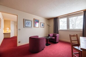 A room with a red carpet, a wooden chair, a table, a sofa, and a window with curtains. - Crystal Hôtel Courchevel (Les Allues)