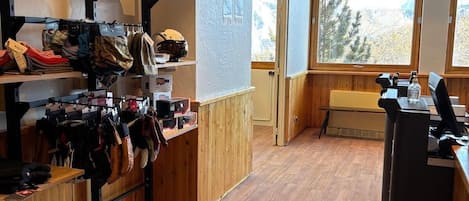 Quarto com piso de madeira, uma escrivaninha com computador, prateleiras com capacetes e luvas, e uma janela com vista para árvores.
