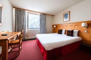 Ett hotellrum med en säng, ett skrivbord och en stol. Det finns ett fönster med gardiner, en träpanel vid sänggaveln och en vägglampa.