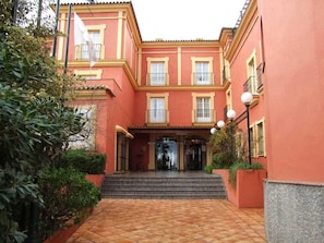 Un edificio con fachada roja, una entrada central con escalones y un patio adoquinado.